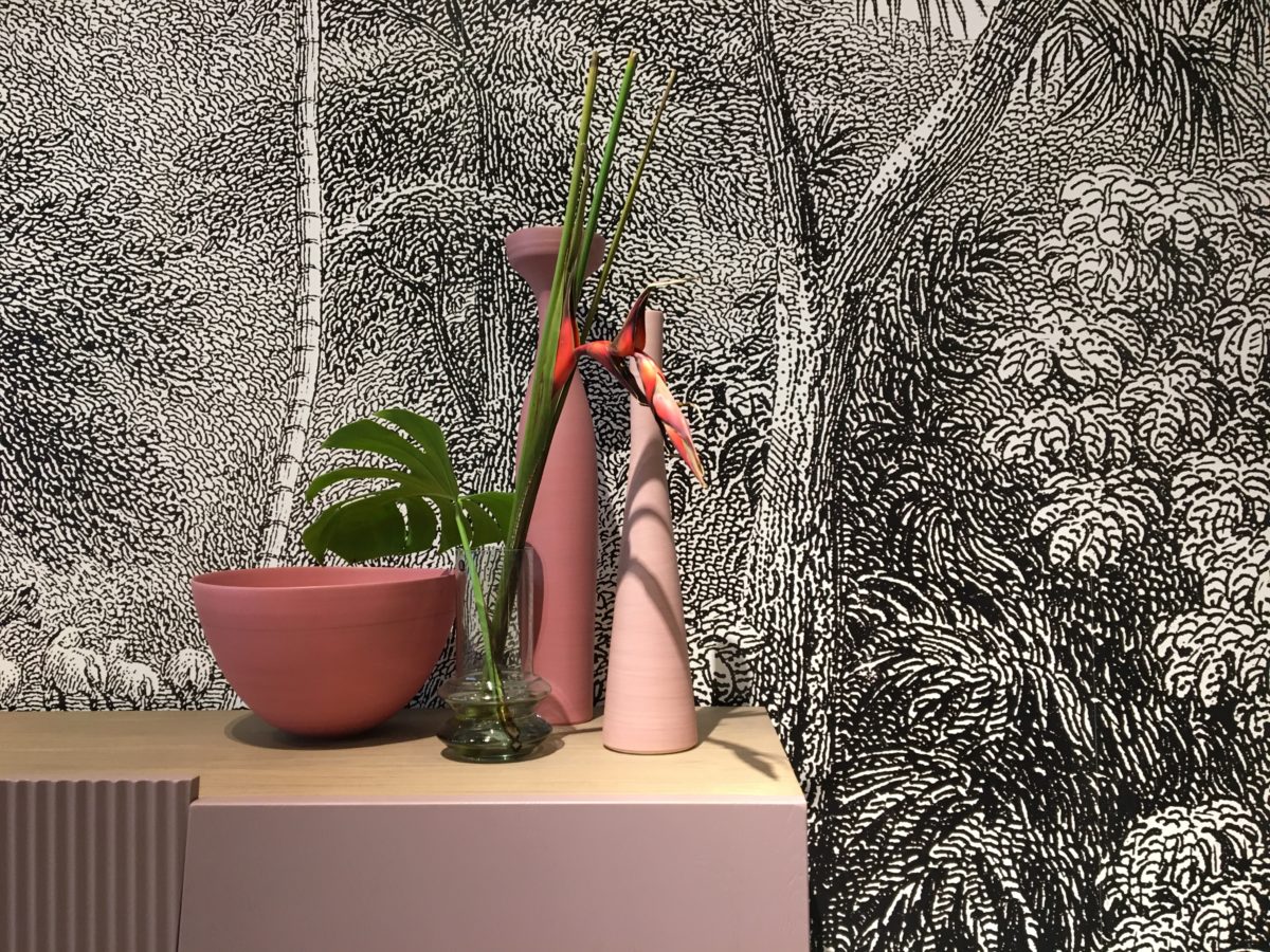 2 vases et une poterie de couleur rose nuancé trônent sur ue buffet laqué rose. Derrière un papier peint esprit jungle en noir et blanc