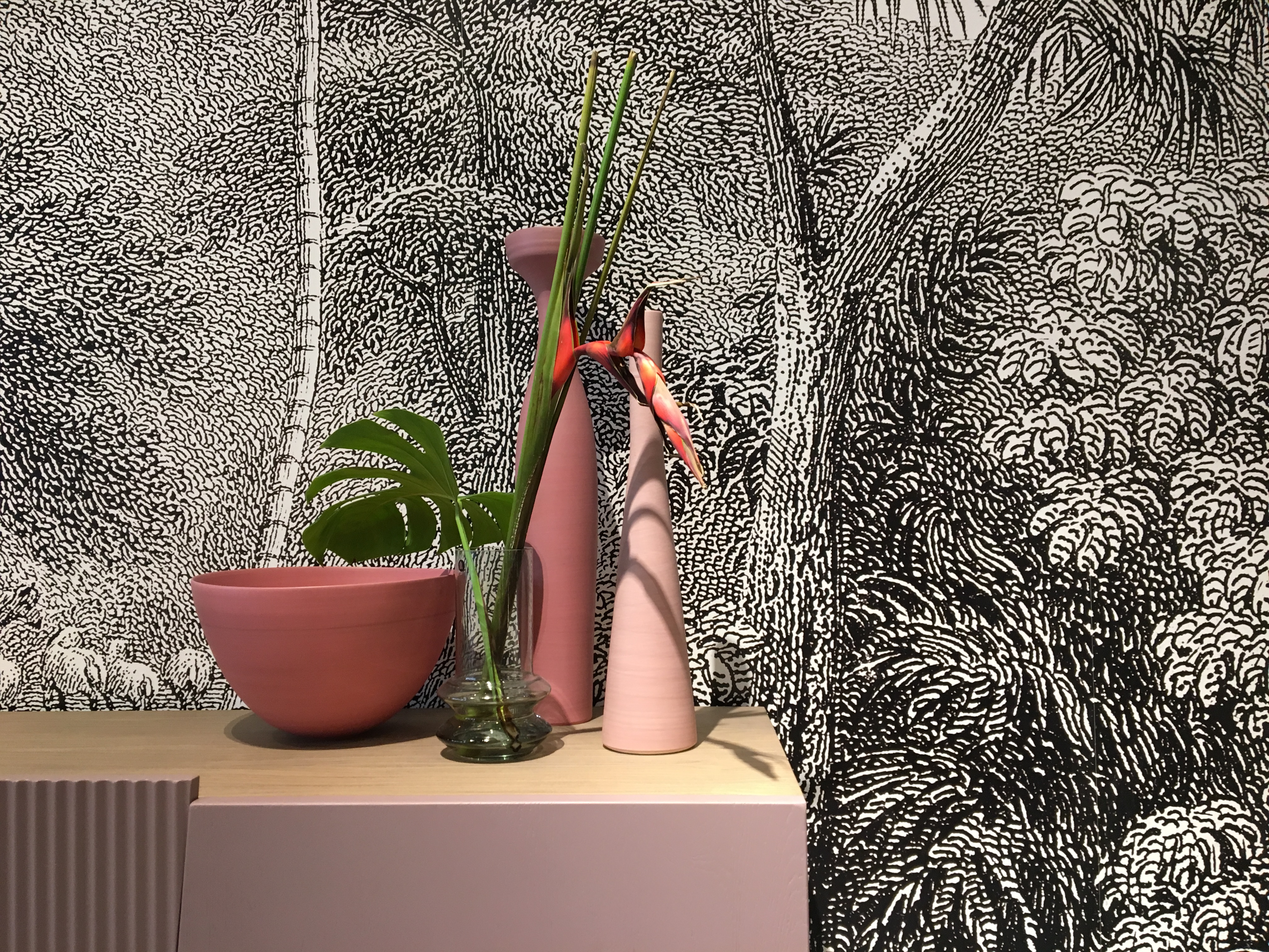 2 vases et une poterie de couleur rose nuancé trônent sur ue buffet laqué rose. Derrière un papier peint esprit jungle en noir et blanc