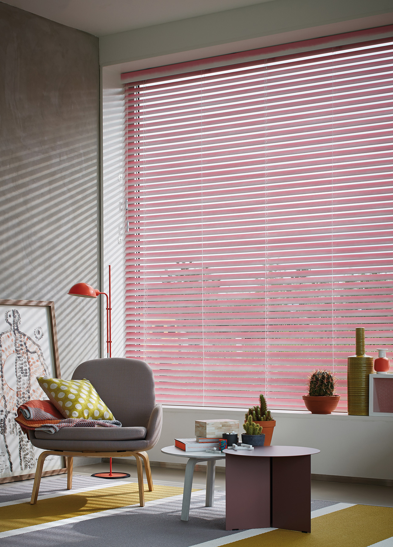 Un grand store vénitein de couleur rose équipe la baie vitrée d'un salon cosy. C'est une création Luxaflex.