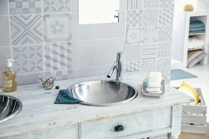 Dans un coin salle de bains une vasque en inox est inscrutée dans un meuble en bois vielli. En crédence, des lambris muraux PVC Grosfillex imitation carreaux ciment.