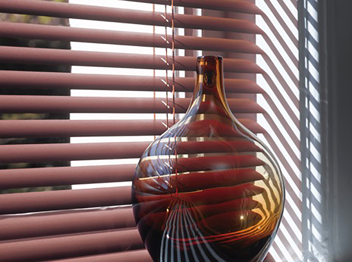 une fenêtre est fermée par un stores vénitens à lames fines laquées couleur vieux rose. Devant un vase transparent et bombé, d'une belle teinte brune.