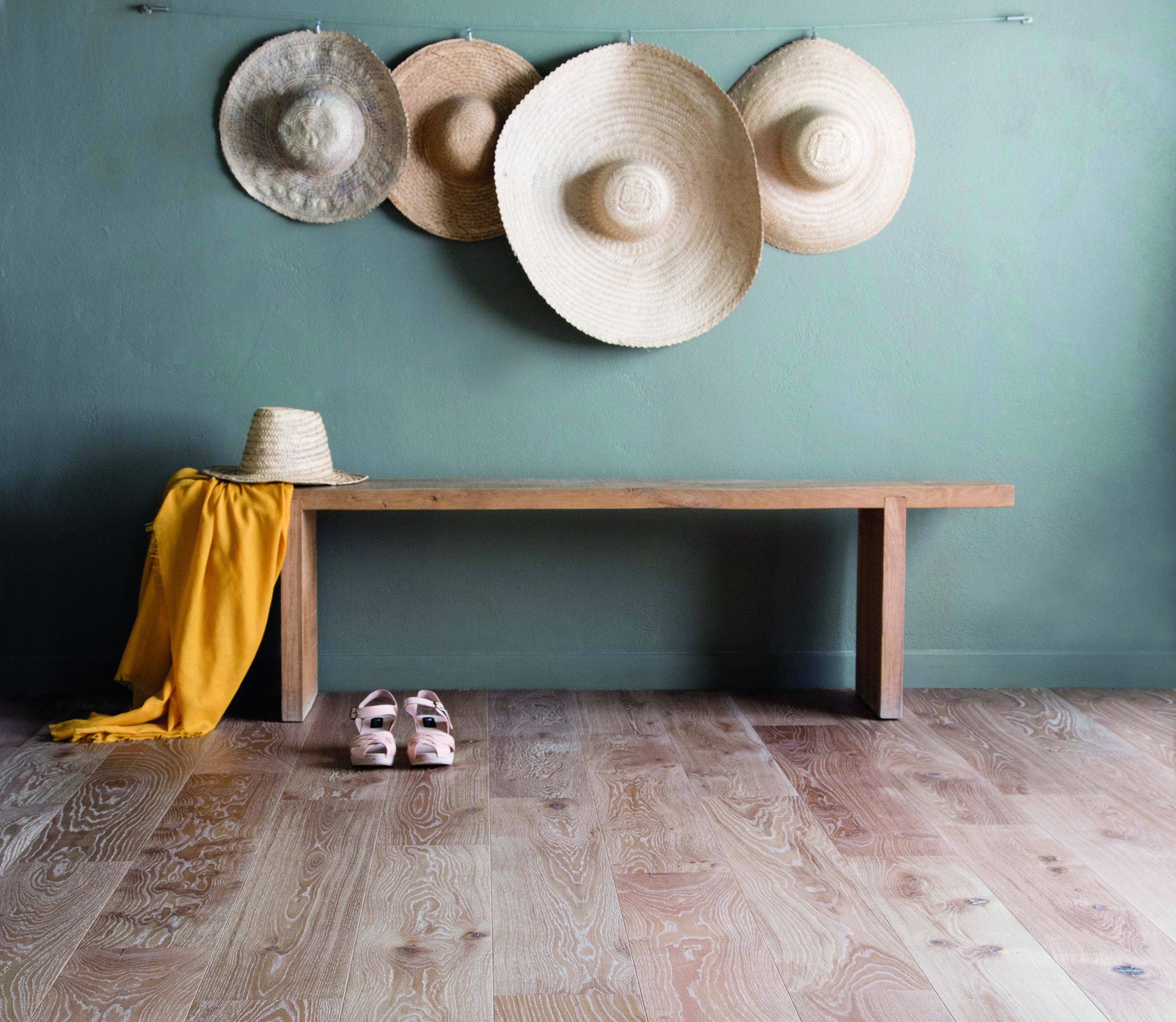 dans une entreé, un banc en bois sur un parquet clair. Le mur peint en vert d'eau accueille 4 grands chapeaux de paille accrochés comme des tabelaux. Un air de printemps