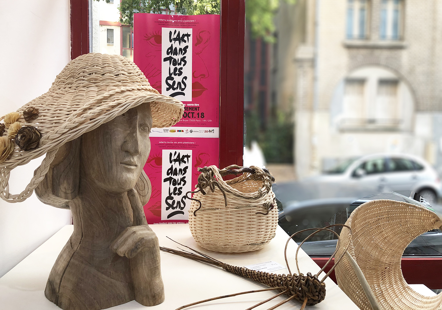 une tête de femme chapeautée est sculptée dans du bois. À cpoté des sculptres en vannerie. Le tout dans une vitrine sur laquelle est affihcée "lart dans tous les sens"