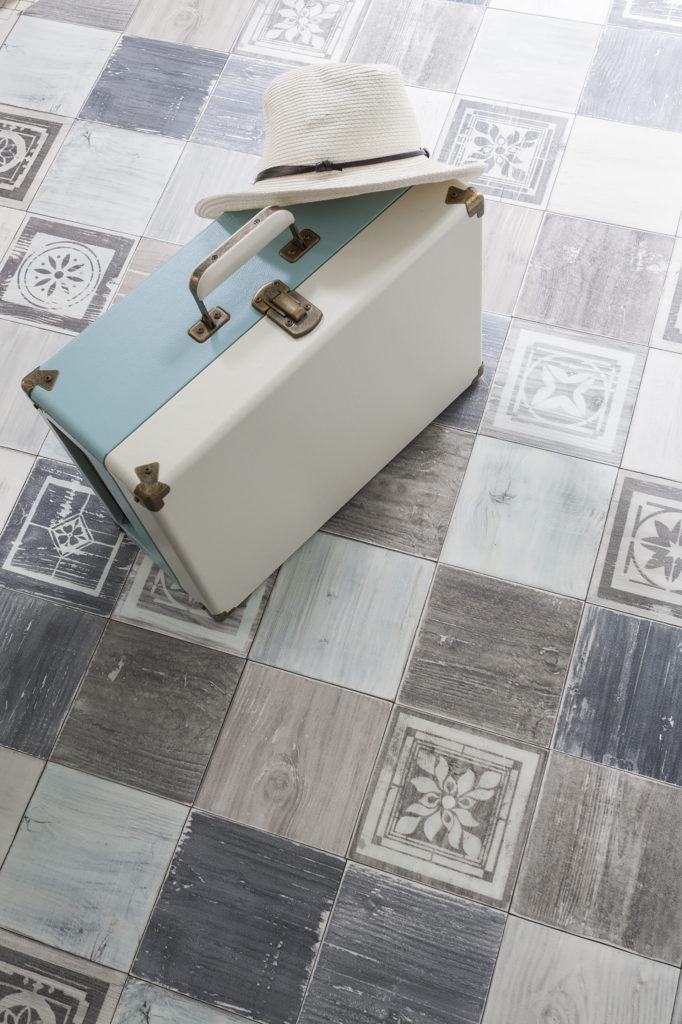 une valise vintage et un panama sont posé surun sol pvc effet carreaux ciment dans les tons bruns, gris et bleu passés.