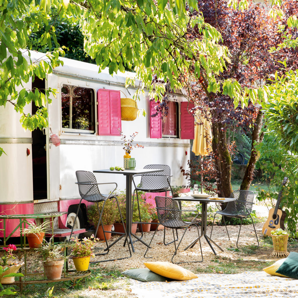 Ambiance bohême avec les chaises Ramatuelle 73' en version hautes et standard. Elles sont posées devant une roulotte aménagée aux volets rose vif, au fond d'un jardin ensoleillé.