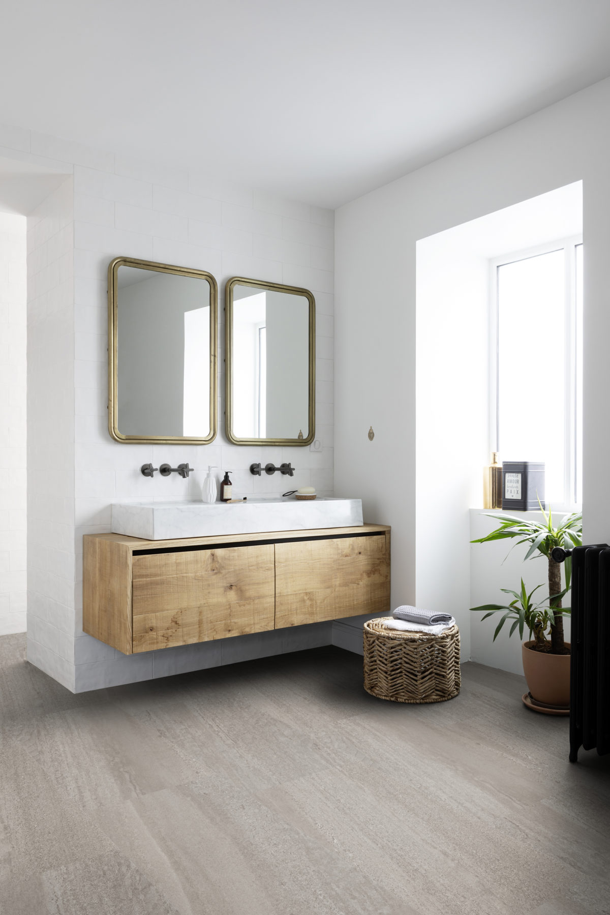 une salle de bain luineuse : murs blancs, meuble en bois qui supporte deux vasques blanches, miroirs jumaux anciens avec cadre couleur or. Au sol un joli sol vinyle Gerflor
