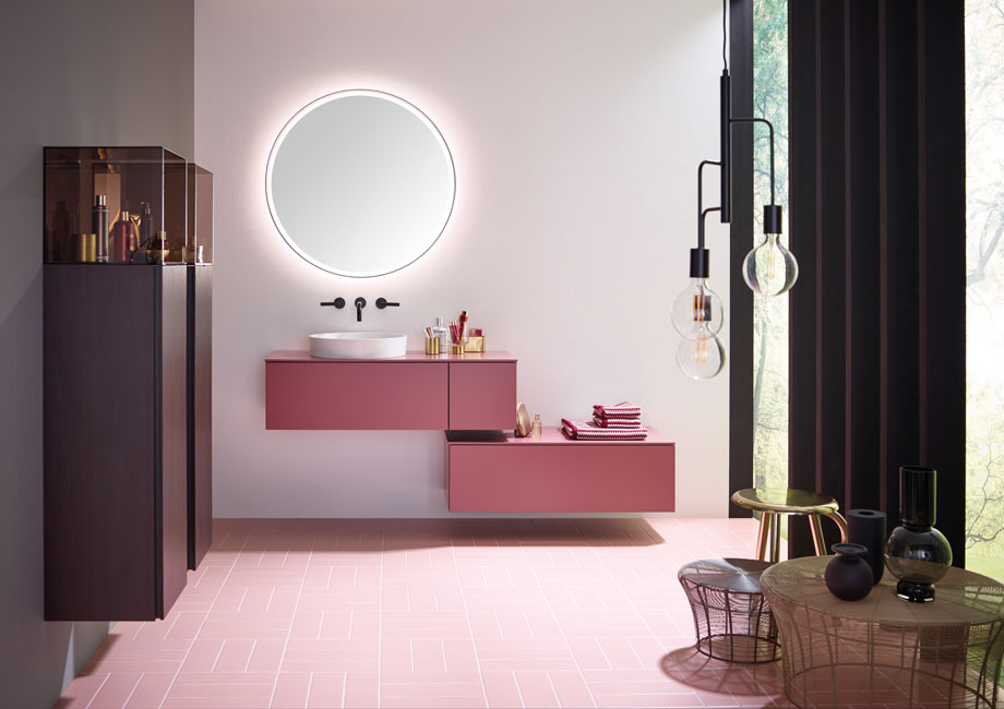 Salle de bains rose. Deux meubles suspendus rose framboise sont surmonté d'un miroir rond géant pour une salle de basin à la fois futuriste et délicate.