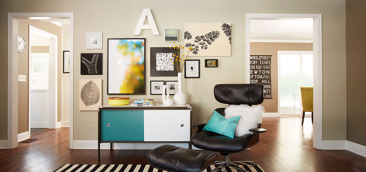 Sur le mur beige d'un coin salon, sont disposés différents cadres décoratifs contenant photos, dessins, lettrages, fleurs séchées. Dessous une enfilade de style scandinave bicolore, un fauteuol en cuir noir de Eames et au sol un petit tapis rayé noir et blanc