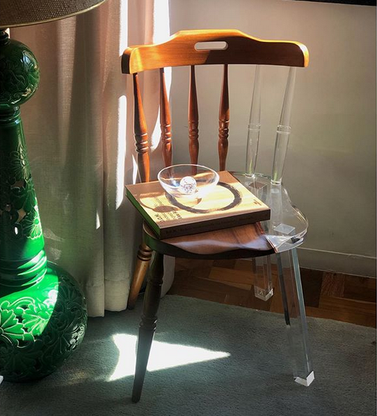 Une chaise rustique en bois dont la partie manquante a été remplacée par une pièce en acrylique transparent. Dessus est posé un petit plateau en bois