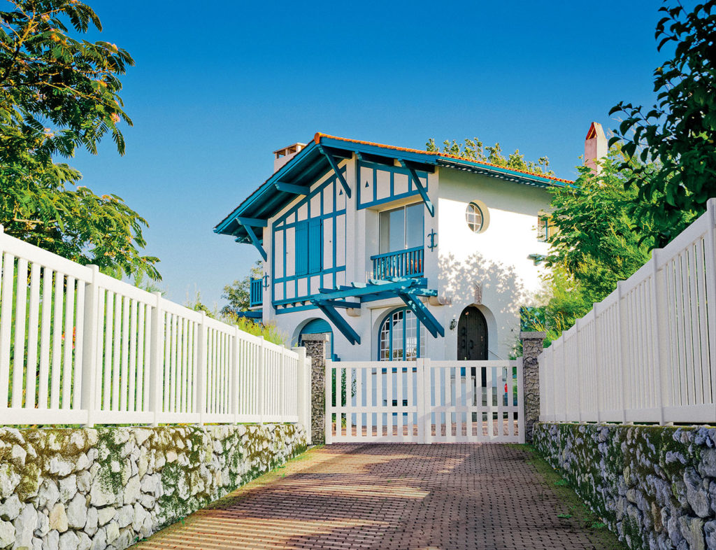 Une maison de style basque aux volets bleus. Elle est fermée par un portail blanc Préfalu