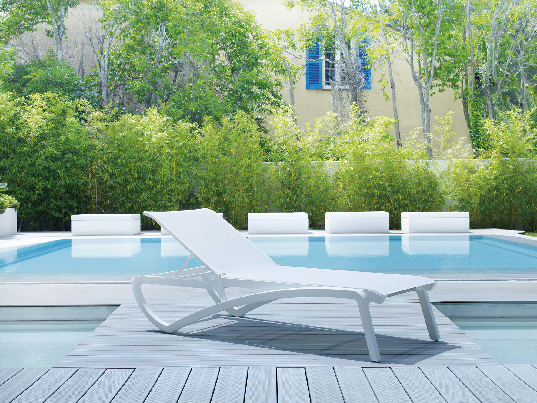 un bains de soleil blanc est posé devant une piscine bleu azur. a l'arrire plan on devine une bastide provençale