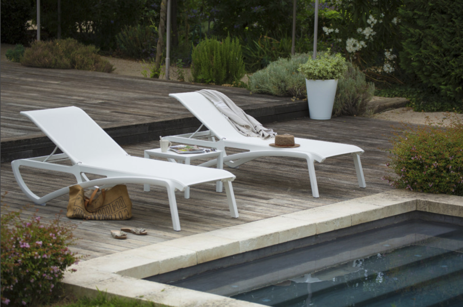 Deux bains de soleil blancs au bord d'une piscine. Un chapeau de paille et un cabas sont posés